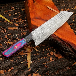 May include: A Damascus steel chef's knife with a vibrant handle. The blade has a textured pattern, and the handle features pink, blue, and gold accents. The knife rests on a piece of wood, showcasing its craftsmanship.