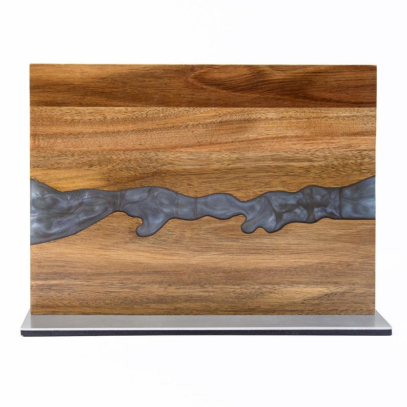 K&ouml;nnte beinhalten: Holzschneidebrett mit einem Fluss aus dunkelgrauem Harz. Das Brett ist rechteckig und hat ein nat&uuml;rliches Holzmaserungsmuster. Das Harz verl&auml;uft horizontal &uuml;ber das Brett. Es ist auf einem silbernen Sockel montiert.