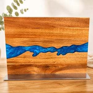 K&ouml;nnte beinhalten: Ein Holzblock mit einem Fluss aus blauem Kunstharz. Das Holz hat einen warmen, rotbraunen Farbton, und das Kunstharz ist ein leuchtendes, gl&auml;nzendes Blau. Der Block ist auf einem silberfarbenen Metallsockel montiert.