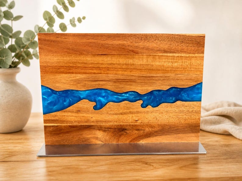 K&ouml;nnte beinhalten: Ein Holzblock mit einem Fluss aus blauem Kunstharz. Das Holz hat einen warmen, rotbraunen Farbton, und das Kunstharz ist ein leuchtendes, gl&auml;nzendes Blau. Der Block ist auf einem silberfarbenen Metallsockel montiert.