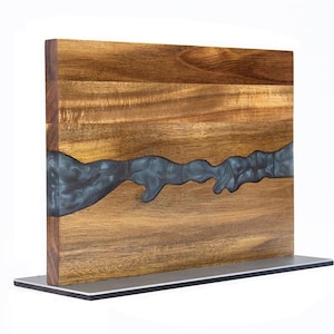 K&ouml;nnte beinhalten: Ein Holzschneidebrett mit einem dunkelgrauen Epoxidharz-Flussdesign. Das Brett besteht aus hellbraunem Holz und steht auf einem silbernen Metallsockel. Der Epoxidharz-Fluss verl&auml;uft &uuml;ber die Mitte des Bretts.