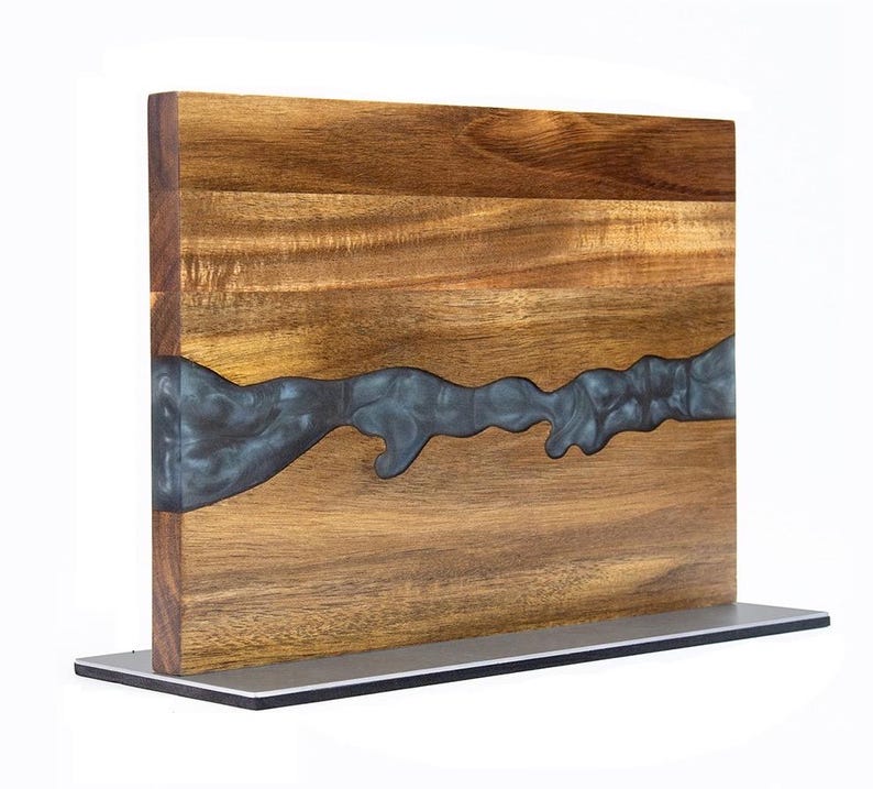 K&ouml;nnte beinhalten: Ein Holzschneidebrett mit einem dunkelgrauen Epoxidharz-Flussdesign. Das Brett besteht aus hellbraunem Holz und steht auf einem silbernen Metallsockel. Der Epoxidharz-Fluss verl&auml;uft &uuml;ber die Mitte des Bretts.