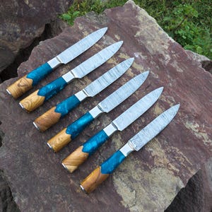 May include: Six Damascus steel knives with blue resin and wood handles are arranged on a large rock. The blades have a distinctive pattern. The handles are a combination of wood and blue resin, with a silver bolster.