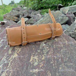 May include: A brown leather roll with white stitching and silver buckles. The roll is closed and sitting on a pile of rocks.
