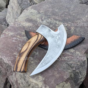 May include: A silver axe with a wooden handle and a leather sheath. The axe head has a decorative design with a compass and a dragon. The axe is engraved with the text "RFPMI FARXM".