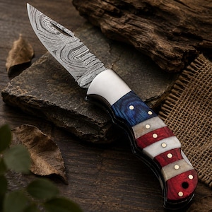 May include: A folding knife with a patterned steel blade and a handle featuring blue, white, and red segments, reminiscent of a flag design. The handle is accented with small, gold-colored rivets. The knife is displayed on a dark, textured surface.