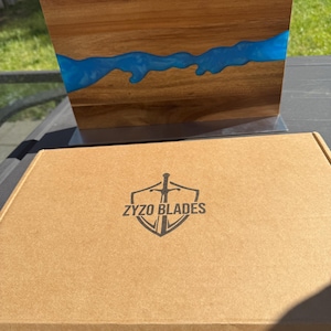 K&ouml;nnte beinhalten: Ein Holzschneidebrett mit einem blauen Harzfluss-Design. Das Brett wird auf einem Metallst&auml;nder pr&auml;sentiert. Eine Pappschachtel mit dem Text "ZYZO BLADES" und einem Schwertlogo befindet sich vor dem Brett.