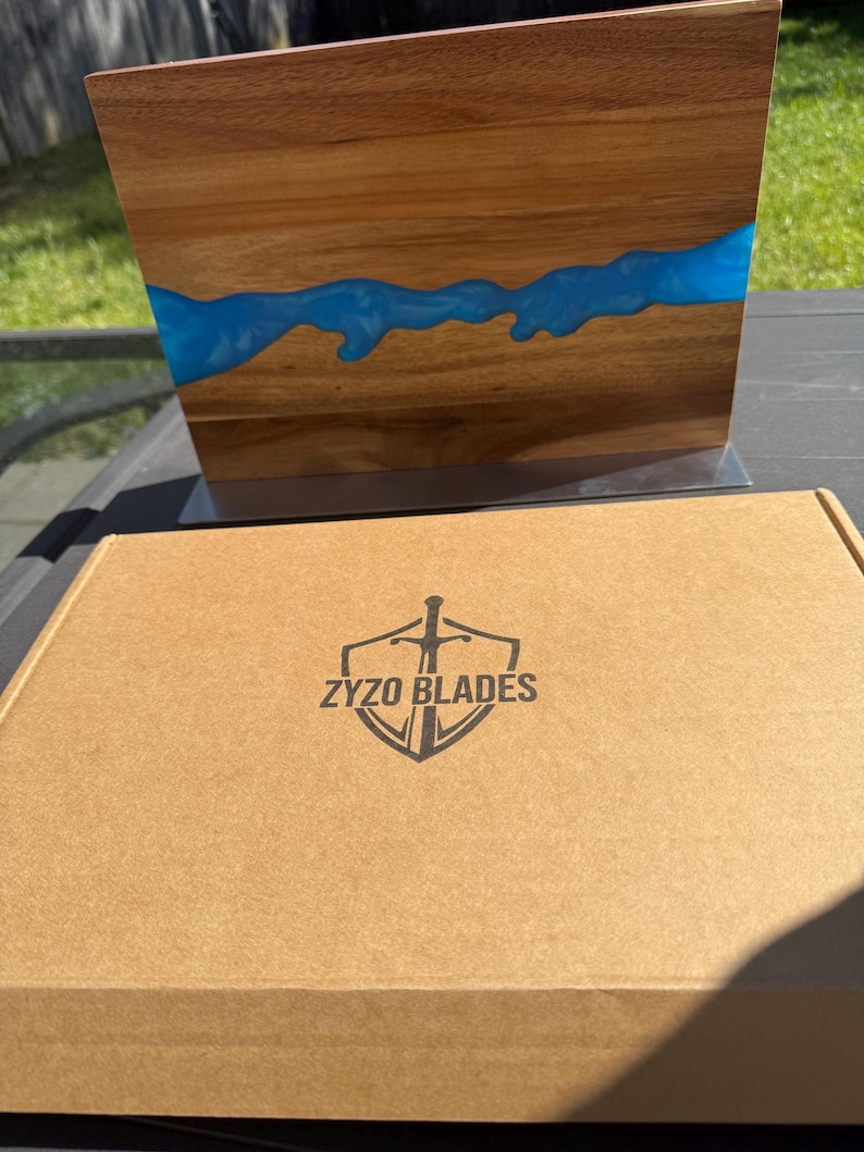 K&ouml;nnte beinhalten: Ein Holzschneidebrett mit einem blauen Harzfluss-Design. Das Brett wird auf einem Metallst&auml;nder pr&auml;sentiert. Eine Pappschachtel mit dem Text "ZYZO BLADES" und einem Schwertlogo befindet sich vor dem Brett.