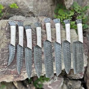 May include: Eight kitchen knives with white bone handles and Damascus steel blades. The knives are arranged in a row on a rocky surface.
