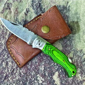 May include: A green and silver pocket knife with a wood handle and a leather sheath. The blade has a Damascus pattern.