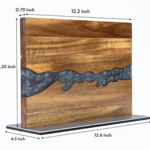 K&ouml;nnte beinhalten: Holzschneidebrett mit einem Flussdesign aus dunkelblauem Harz. Das Brett ist 33,5 cm lang, 23,5 cm hoch und 2 cm dick. Es steht auf einem Metallsockel, der 34,5 cm lang und 11,4 cm breit ist. Das Holz hat eine nat&uuml;rliche Maserung.