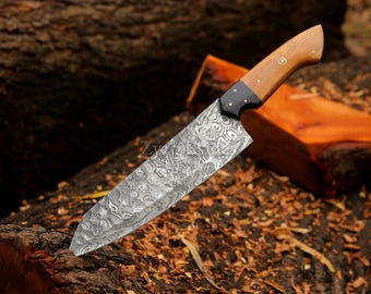 May include: A Damascus steel chef's knife with a wooden handle. The blade has a detailed pattern, and the handle is a light brown color with black accents. The knife is resting on a bed of wood shavings and logs.