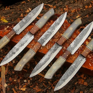 May include: Six handmade knives with textured blades and wooden handles. The handles combine light grey, dark brown, and gold accents. The knives are displayed on a wooden surface. The blades have a rustic finish.