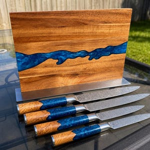 K&ouml;nnte beinhalten: Ein Messerblock aus Holz mit einem blauen Harzfluss-Design, der vier Messer enth&auml;lt. Die Messer haben gemusterte Klingen, blaue und holzfarbene Griffe und einen Edelstahl-Handschutz. Der Block besteht aus Holz und hat einen Edelstahlfu&szlig;.