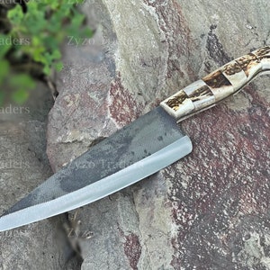 May include: A black chef's knife with a textured blade and a brown and white antler handle. The knife is lying on a gray rock.