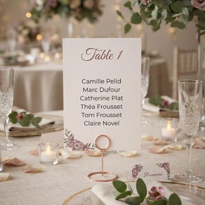 May include: A rectangular white table number card labeled Table 1 with a list of names including Camille Pelid, Marc Dufour, Catherine Plat, Théa Frousset, Tom Frousset, and Claire Novel, featuring floral accents and a rose gold circular stand.