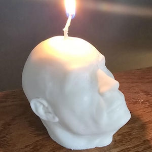 May include: A white candle shaped like a human head with a lit wick on top.