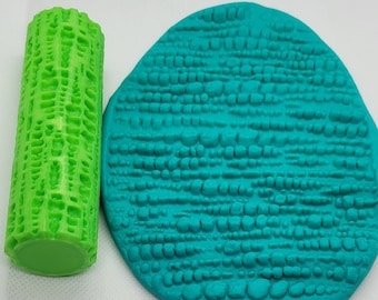 May include: A green plastic roller with a textured surface and a turquoise blue piece of clay with the same texture. The roller is used to create the texture on the clay.