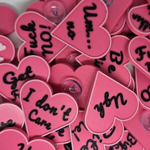 Puede incluir: Un conjunto de pegatinas rosas en forma de corazón con texto negro y ventosas transparentes. Las pegatinas presentan frases como "Te amo", "Fuck no!" y "Me da igual". Los corazones están esparcidos sobre una superficie blanca.