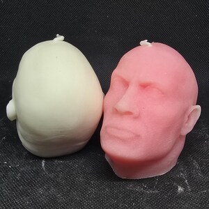 May include: Two head-shaped candles, one white and one pink. The pink candle has a detailed face sculpted on it.