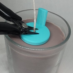 Puede incluir: Primer plano de un par de cortadores de alambre negros recortando una mecha blanca que se enhebra a través de un portavelas azul turquesa. El portavelas está sentado encima de un frasco de vidrio lleno de cera de vela marrón claro.