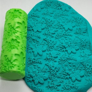 Puede incluir: Un rodillo verde con un diseño floral se utiliza para crear una impresión en una superficie azul turquesa. El rodillo está hecho de plástico y tiene una superficie texturizada. La superficie está hecha de un material suave, posiblemente arcilla o masa.