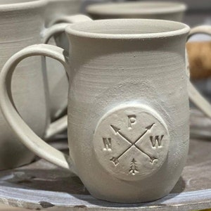 Puede incluir: Una taza de cerámica gris claro con un diseño de brújula que presenta las letras N, W, P y E. La taza tiene un asa y está sentada sobre un soporte para pasteles blanco.