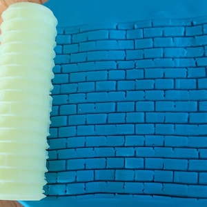 Może przedstawiać: Jasnozielony wałek do ciasta z fakturowaną powierzchnią służy do tworzenia wzoru ceglanego na niebieskiej powierzchni z gliny.