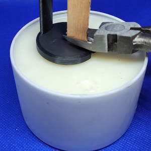 May include: A white candle jar with a black wick holder and a wooden wick being cut with pliers. The candle jar is on a blue surface.