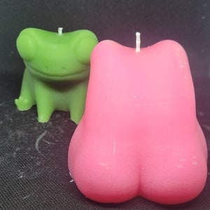 May include: Two novelty candles: a green frog candle and a pink candle shaped like a rounded form. Both candles have white wicks. The frog candle is in a sitting position. The pink candle has a rounded bottom.