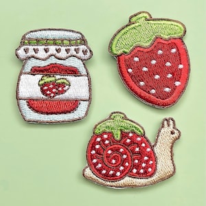 Puede incluir: Tres parches bordados para planchar que representan una fresa, un tarro de mermelada de fresa y un caracol que lleva una fresa a la espalda.