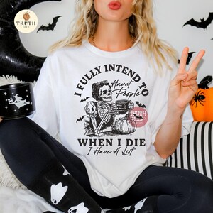 May include: White t-shirt with a Halloween graphic. The design includes a skeleton with a mug, a pumpkin, and the text "I FULLY INTEND TO HAUNT PEOPLE WHEN I DIE I Have A List". The shirt has a crew neck and short sleeves.