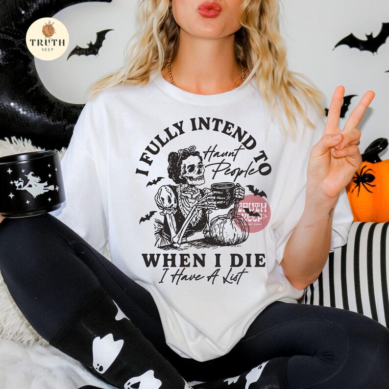May include: White t-shirt with a Halloween graphic. The design includes a skeleton with a mug, a pumpkin, and the text "I FULLY INTEND TO HAUNT PEOPLE WHEN I DIE I Have A List". The shirt has a crew neck and short sleeves.