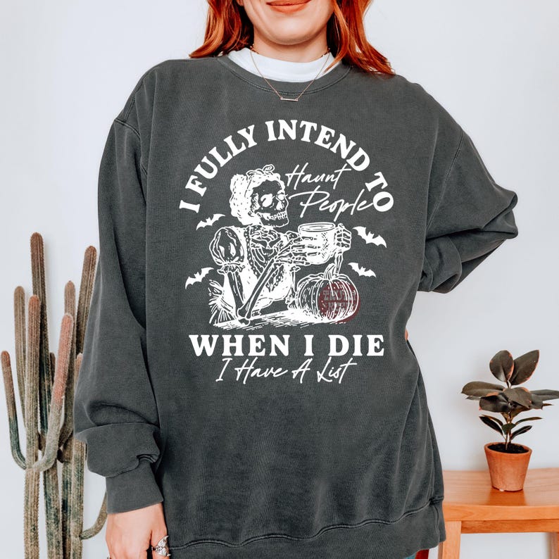 May include: Dark grey sweatshirt featuring a white graphic of a skeleton with a mug and pumpkin. The text says "I FULLY INTEND TO Haunt People WHEN I DIE I Have A List."