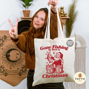Puede incluir: Bolso tote de lona color crema con texto y gr&aacute;fico en rojo. El texto dice "Gone Fishing, Be Back by Christmas". El gr&aacute;fico muestra a Pap&aacute; Noel sosteniendo un pez, con cubos etiquetados "Naughty Fish" y "Nice Fish".