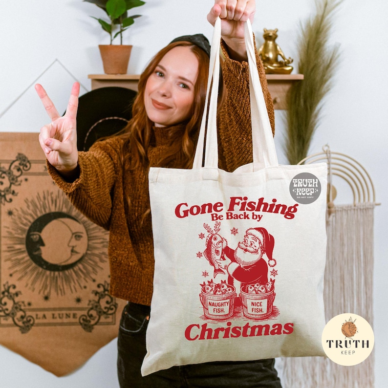 Puede incluir: Bolso tote de lona color crema con texto y gr&aacute;fico en rojo. El texto dice "Gone Fishing, Be Back by Christmas". El gr&aacute;fico muestra a Pap&aacute; Noel sosteniendo un pez, con cubos etiquetados "Naughty Fish" y "Nice Fish".