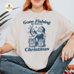 Puede incluir: Camiseta color crema con un gr&aacute;fico azul marino que muestra a Pap&aacute; Noel sosteniendo un pez, con el texto "Gone Fishing, Be Back by Christmas". Debajo, dos cubos con las etiquetas "Naughty Fish" y "Nice Fish".