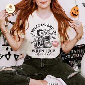 May include: A cream-coloured t-shirt with a Halloween graphic. The design shows a skeleton holding a mug and a pumpkin, with the text "I FULLY INTEND TO HAUNT PEOPLE WHEN I DIE I Have A List."