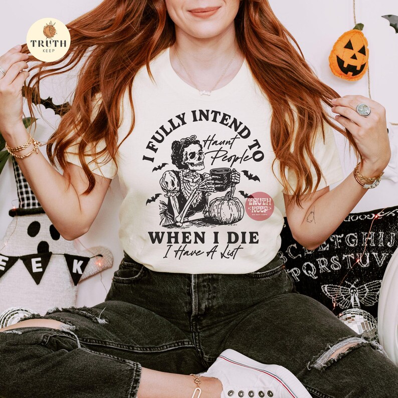 May include: A cream-coloured t-shirt with a Halloween graphic. The design shows a skeleton holding a mug and a pumpkin, with the text "I FULLY INTEND TO HAUNT PEOPLE WHEN I DIE I Have A List."