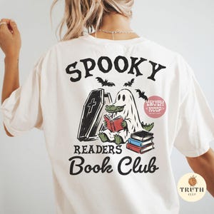 May include: An off-white t-shirt featuring a graphic on the back with the text SPOOKY READERS Book Club. The design shows a green alligator wearing a white ghost sheet, sitting next to a coffin and reading a red book, with a stack of books nearby.