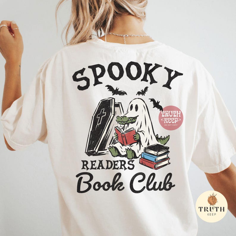 May include: An off-white t-shirt featuring a graphic on the back with the text SPOOKY READERS Book Club. The design shows a green alligator wearing a white ghost sheet, sitting next to a coffin and reading a red book, with a stack of books nearby.