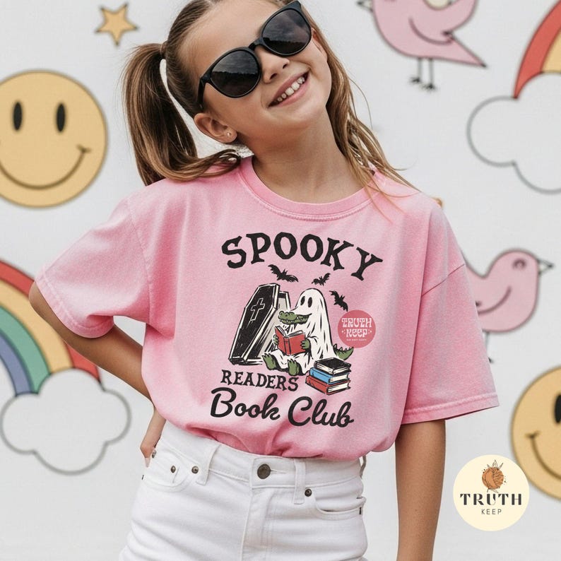 May include: A light pink t-shirt featuring a graphic of a ghost-clad alligator reading a book next to a coffin and a stack of books. The text reads SPOOKY READERS BOOK CLUB and TRUTH KEEP. The shirt has short sleeves and a crew neckline.