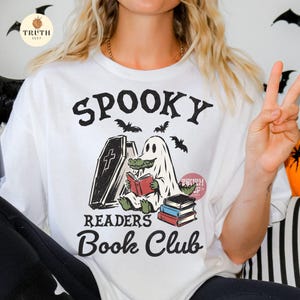 May include: A white t-shirt featuring a graphic of a green alligator wearing a white ghost sheet, sitting next to an open coffin while reading a red book. The design includes a stack of three books, flying bats, and the text SPOOKY READERS BOOK CLUB.