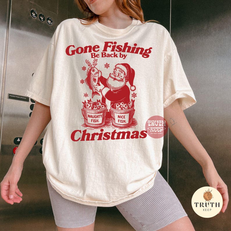 Puede incluir: Camiseta color crema con un gr&aacute;fico rojo que muestra a Pap&aacute; Noel sosteniendo un pez con cuernos. El texto dice "Gone Fishing, Be Back by Christmas". Dos cubos dicen "Naughty Fish" y "Nice Fish".