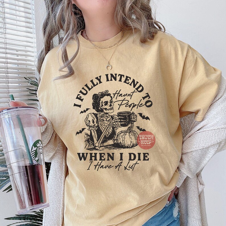 May include: Mustard yellow t-shirt with a Halloween graphic. The design includes a skeleton with a mug and a pumpkin, and the text reads "I Fully Intend to Haunt People When I Die I Have A List."