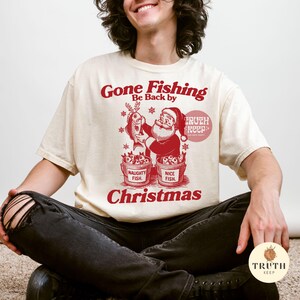Puede incluir: Camiseta color crema con un gr&aacute;fico rojo que presenta a Pap&aacute; Noel sosteniendo un pez, con el texto "Gone Fishing, Be Back by Christmas". Dos cubos etiquetados "Naughty Fish" y "Nice Fish" est&aacute;n debajo.