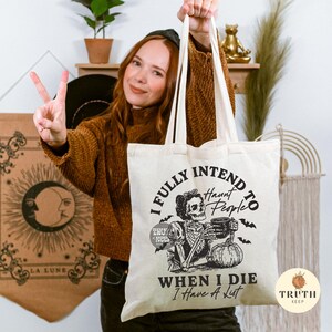 May include: Cream-coloured canvas tote bag featuring a black graphic of a skeleton with a mug and pumpkin. The text reads "I FULLY INTEND to Haunt People WHEN I DIE I Have A List". The bag has two handles and is held up.