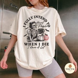 May include: Cream-coloured t-shirt with a black graphic featuring a skeleton holding a mug, a pumpkin, and bats. The text reads "I FULLY INTEND TO HAUNT PEOPLE WHEN I DIE I HAVE A LIST."