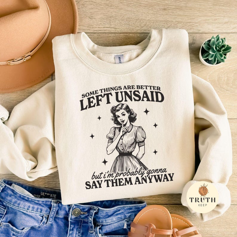 May include: A cream-colored sweatshirt with a black graphic of a woman with her finger to her lips. The text on the sweatshirt reads "Some things are better left unsaid, but I'm probably gonna say them anyway."
