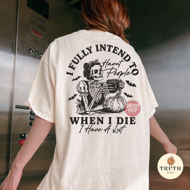 May include: Cream-coloured t-shirt featuring a black skeleton graphic holding a mug and a pumpkin. The text on the shirt reads "I FULLY INTEND TO Haunt People WHEN I DIE I Have A List."